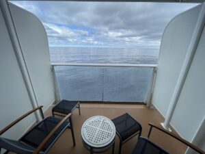 Cruise ship balcony view of white wall partitions on either side with two blue mesh chairs with footstools and one one table in the middle and a view of the water and clouds ahead
