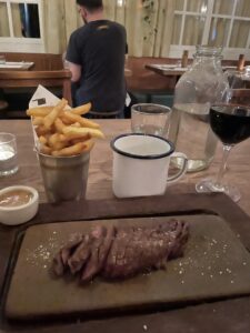 Solo dining at Flat Iron Steak London - steak sliced on a board, cup of french fries, side container of peppercorn sauce and glass of red wine with carafe of water