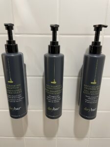 Three wall mounted black bottles of DryBar shower accessories and white tiled wall at Aloft Chelsea NYC